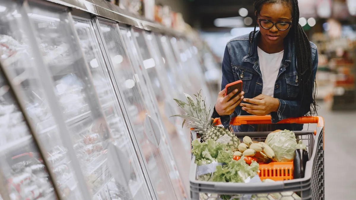 Woman using a smartphone while pushing a shopping cart full of groceries in a supermarket aisle